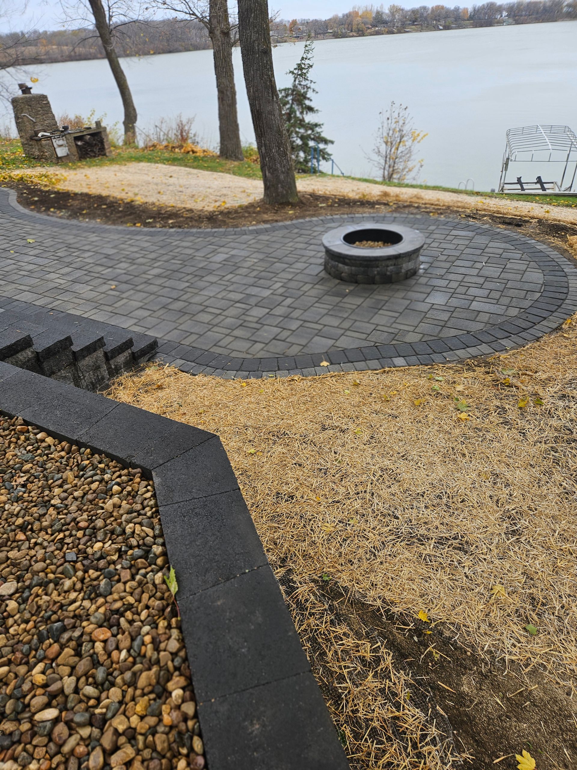 A paved patio with a stone fire pit overlooking a lake, featuring stone retaining walls and decorative gravel pathways.