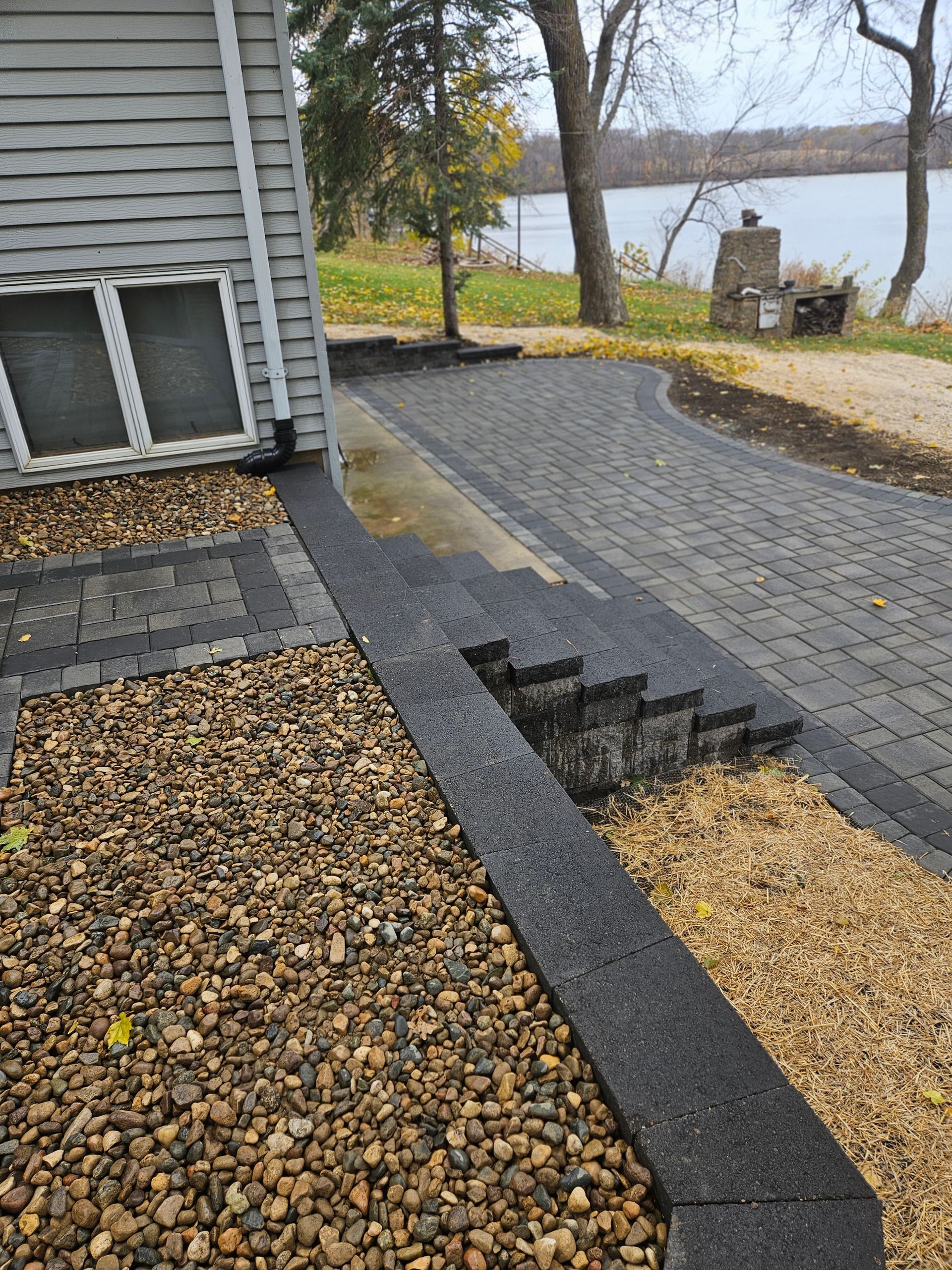 A brick-paved path next to a house with an unfinished section leading to stone stairs, set against a lakeside background.