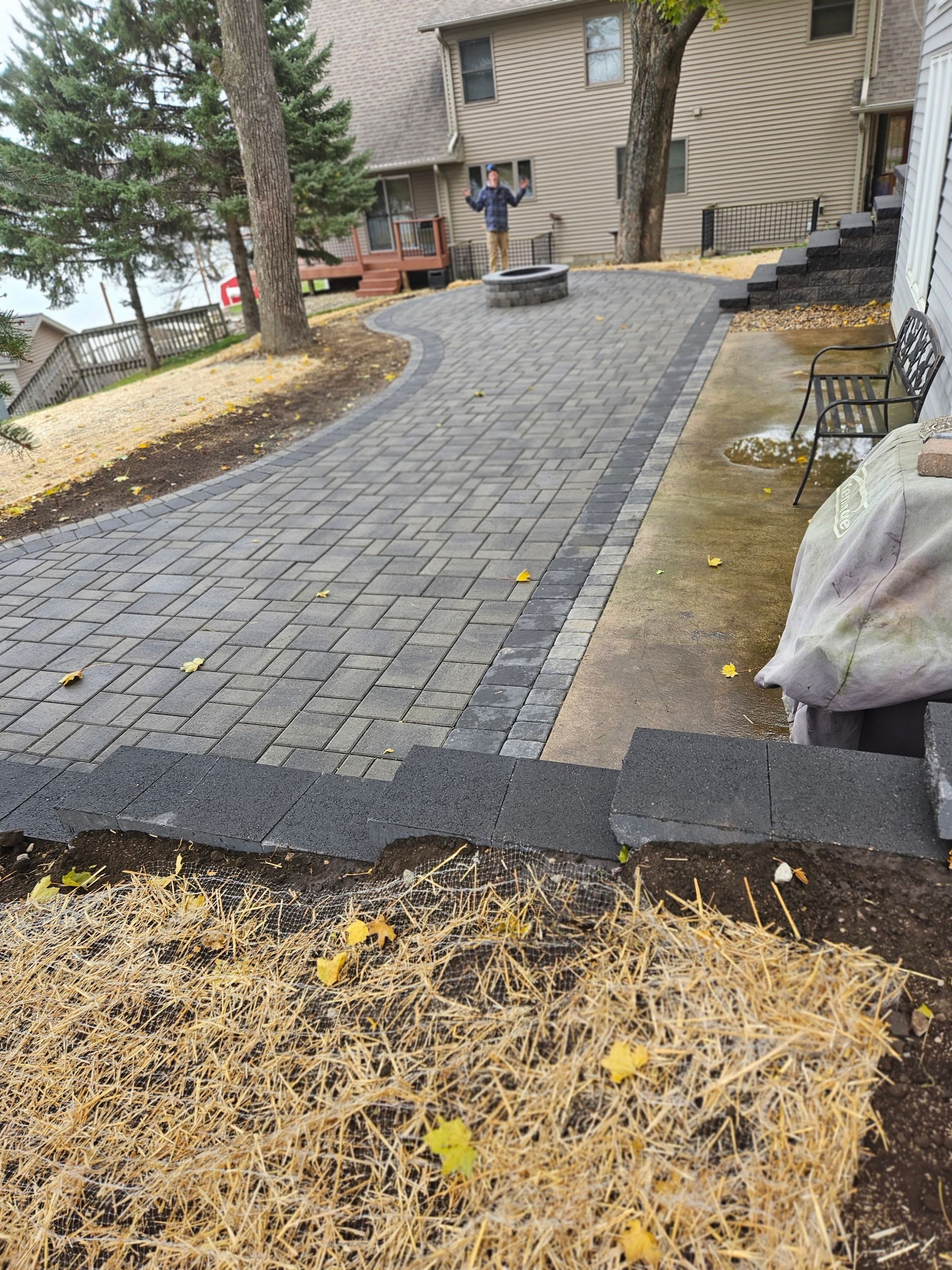 A newly installed gray paver patio with a stone fire pit, tiered wall, and person standing in the background.