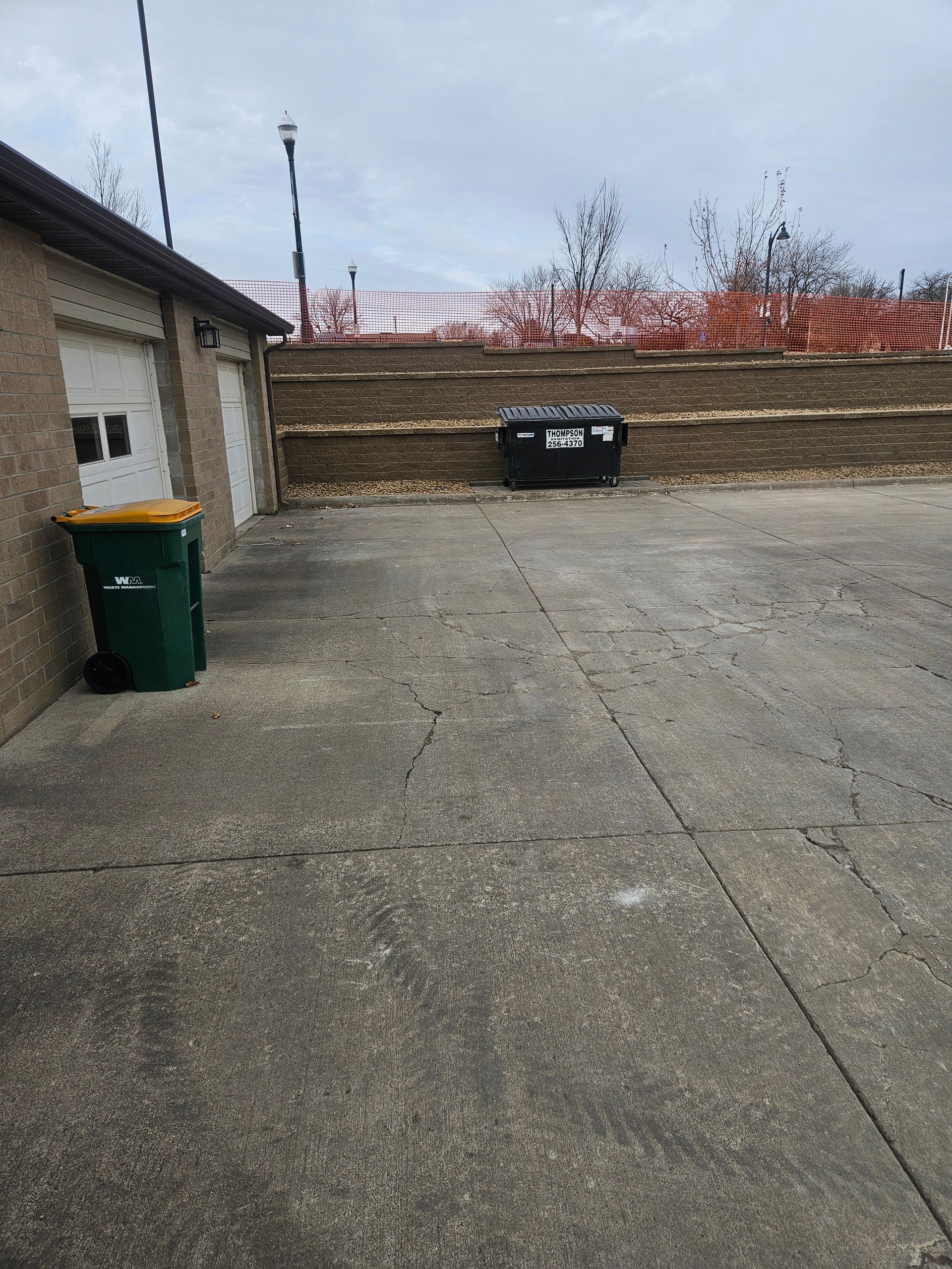 Concrete lot next to a brick building with a green trash bin, a black dumpster, and a fenced-in hillside.