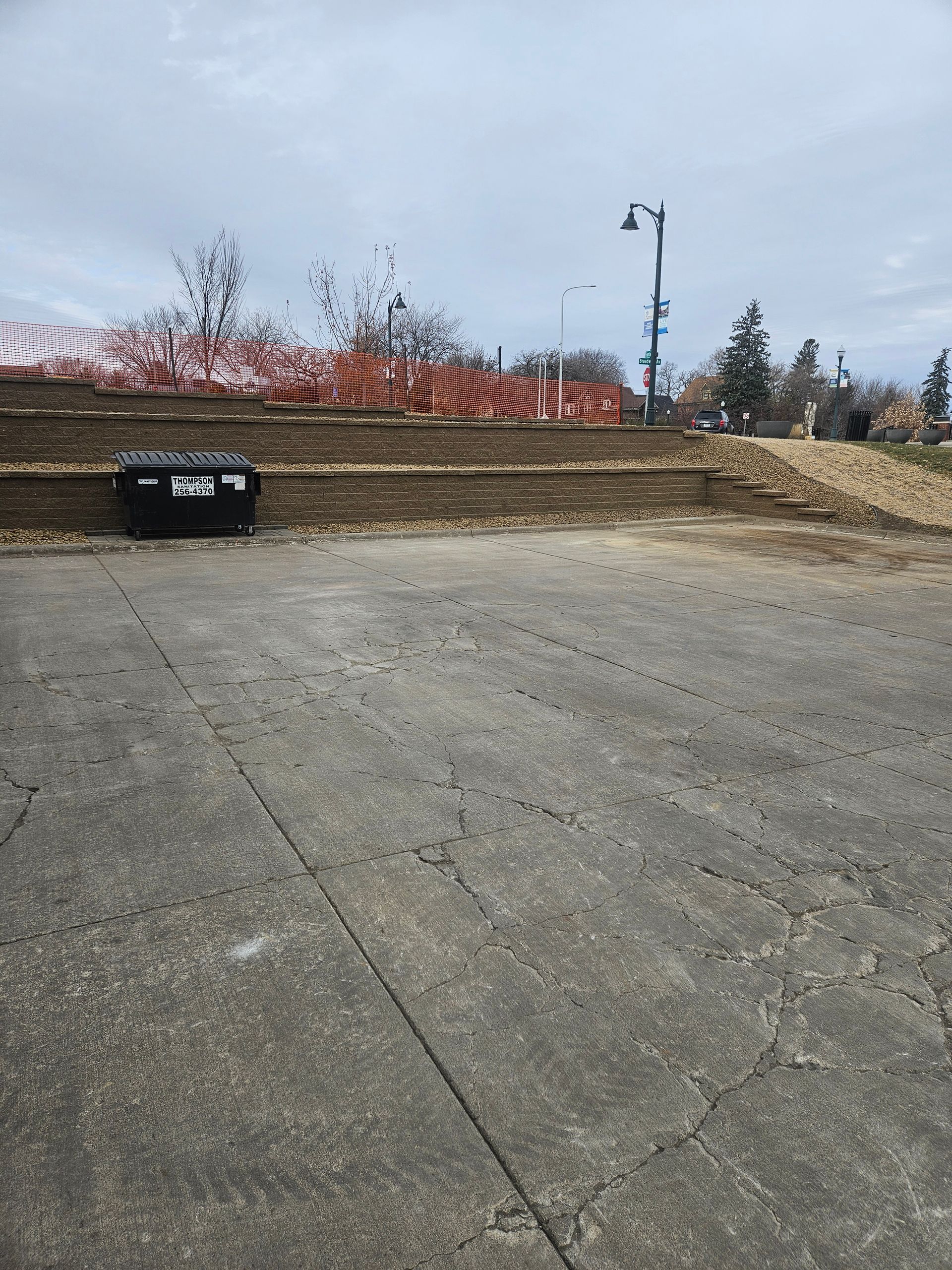 A gravel parking area leading to a terraced dirt hillside with orange construction fencing and a black dumpster.