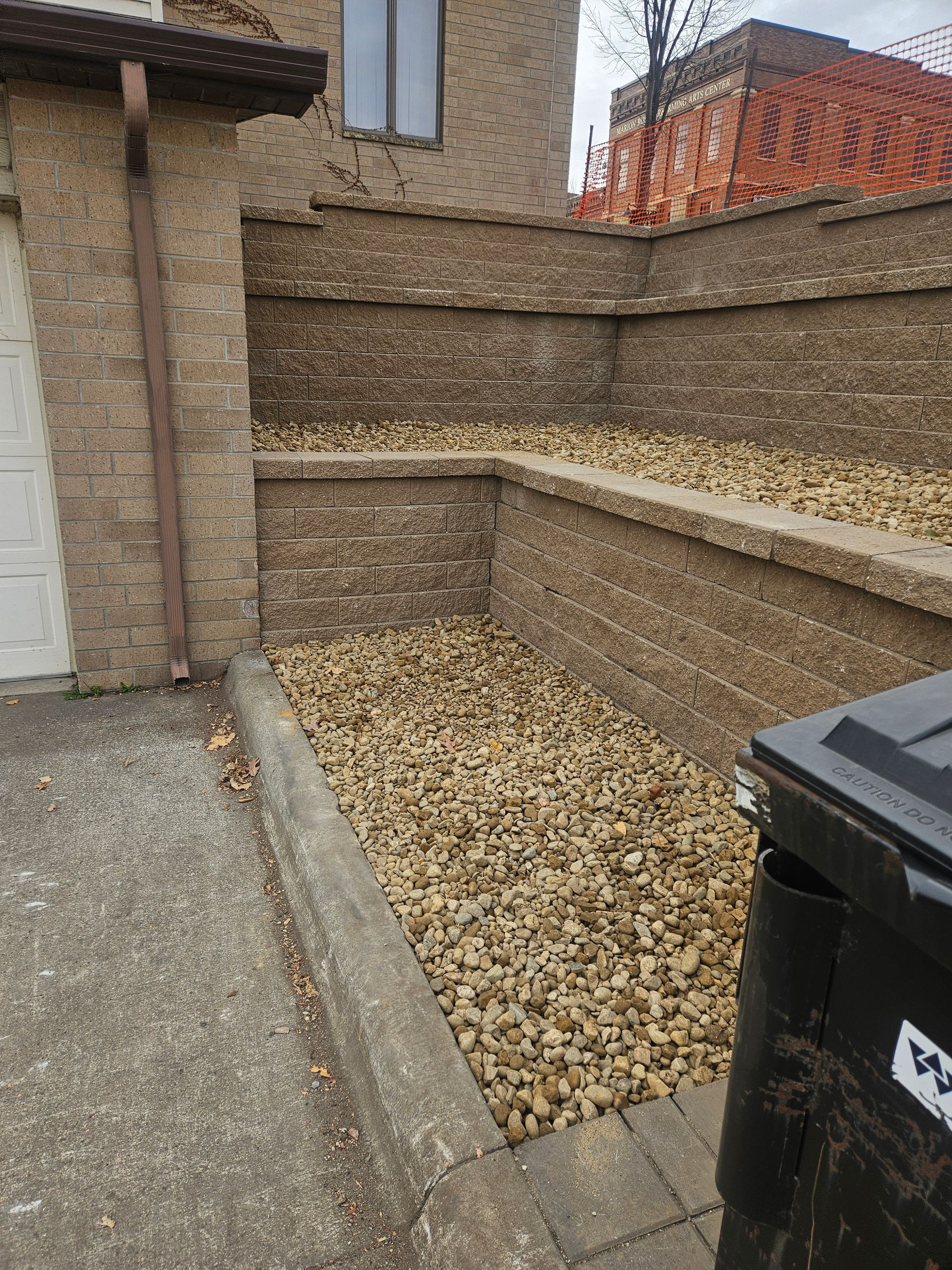 A stone retaining wall with two tiers filled with decorative gravel, adjacent to a concrete driveway and a trash bin.