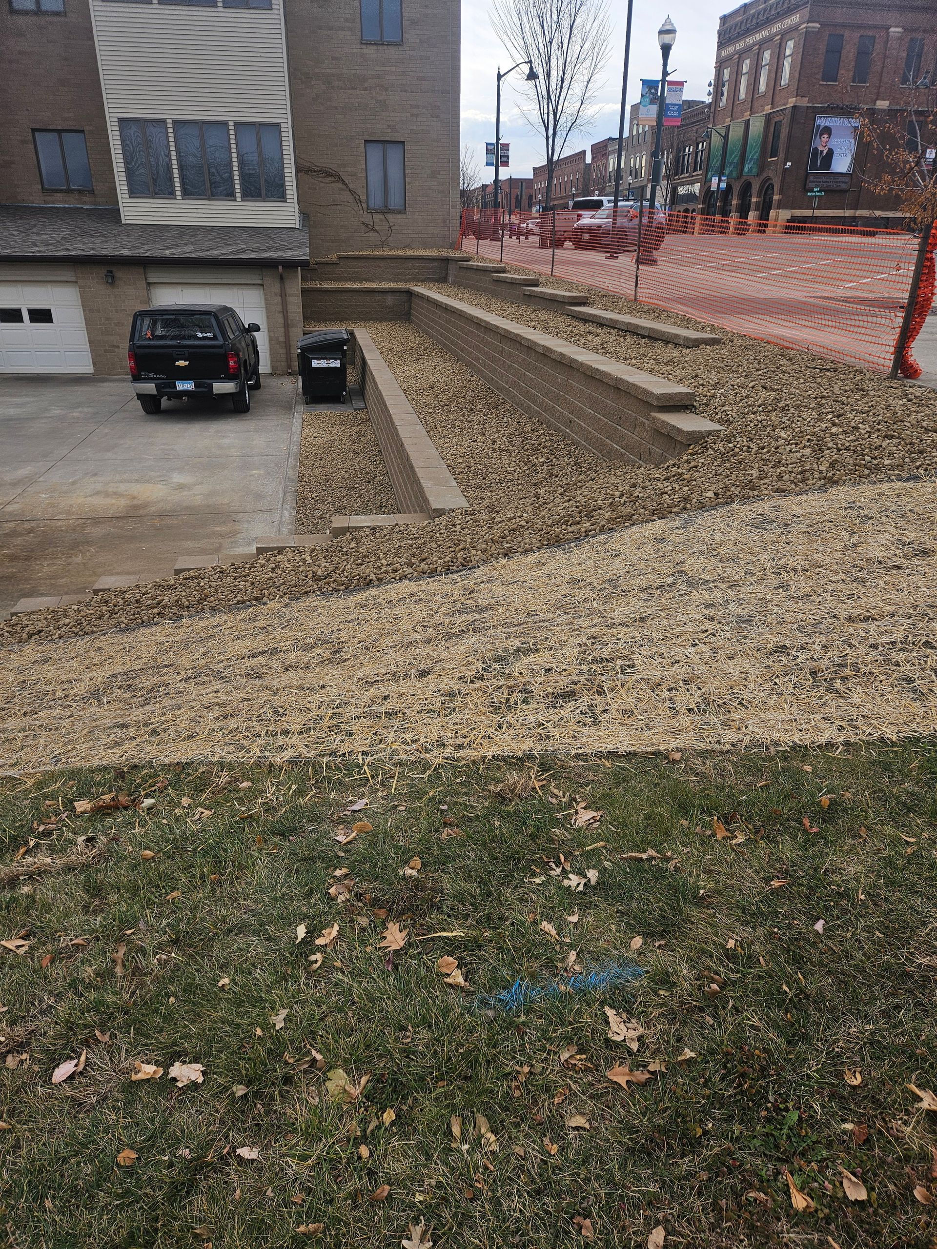 A brick building and retaining wall alongside a gravel driveway with a parked black truck and a street in the background.