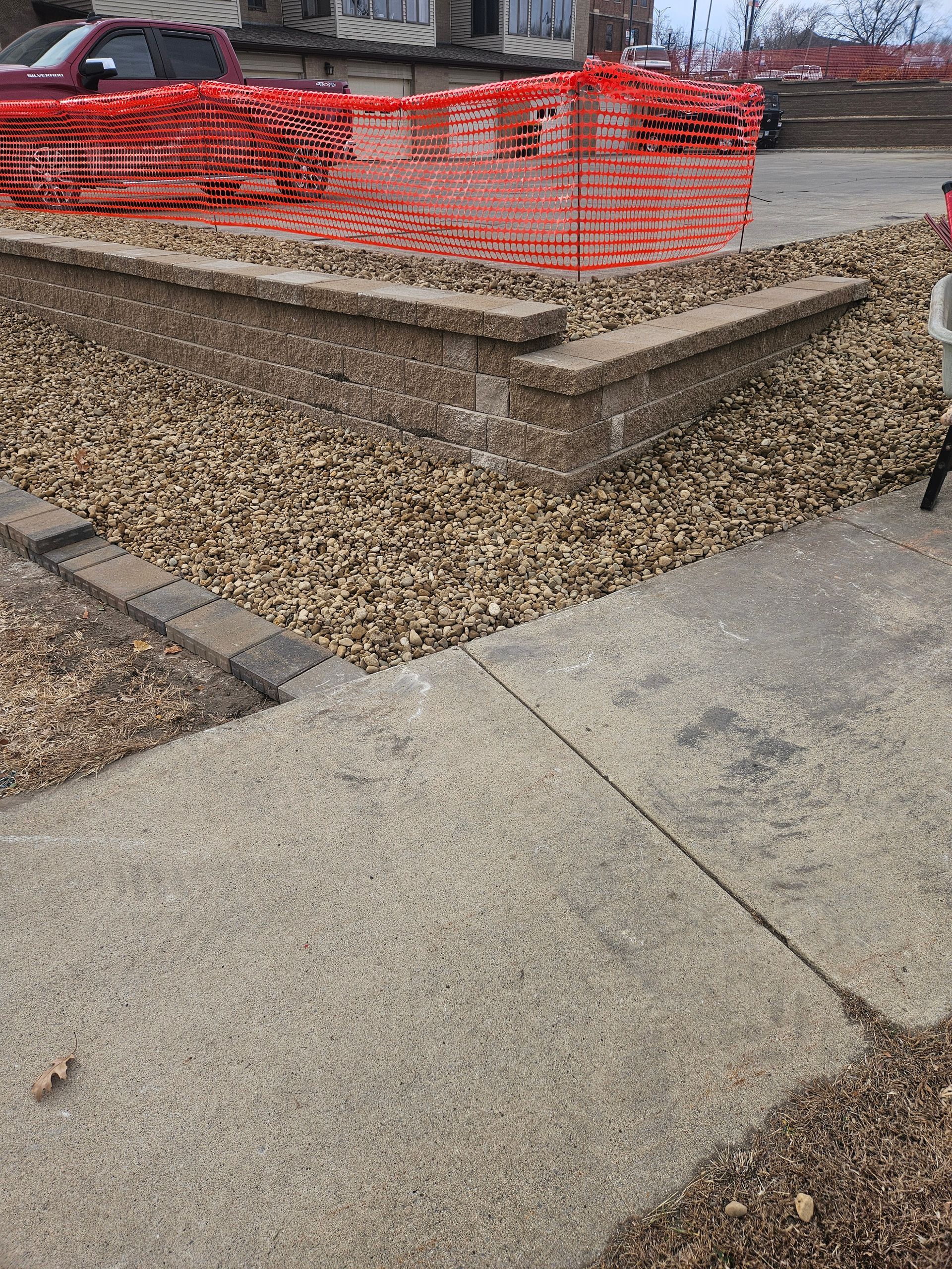A low concrete retaining wall sits behind a gravel bed, bordered by a concrete sidewalk and an orange construction fence.