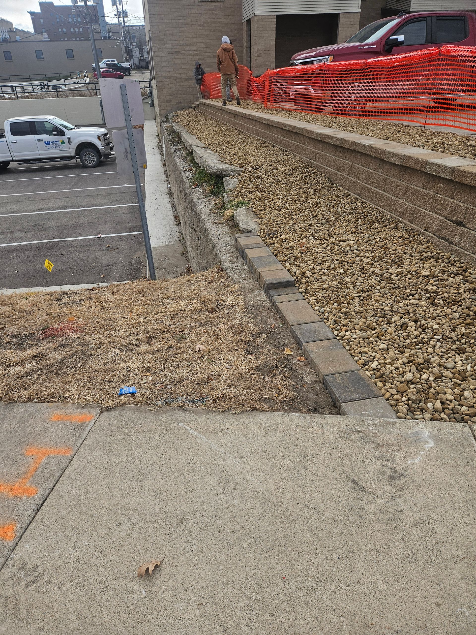 A construction site scene with a worker, an orange safety fence, and a white pickup truck parked on a paved lot.