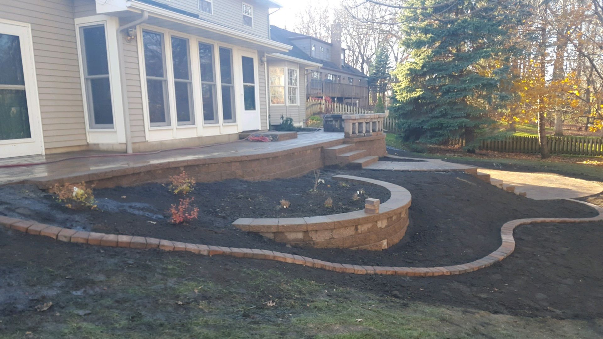 A backyard with a newly installed stone patio, a curved tiered retaining wall, and fresh mulch beds surrounding the house.