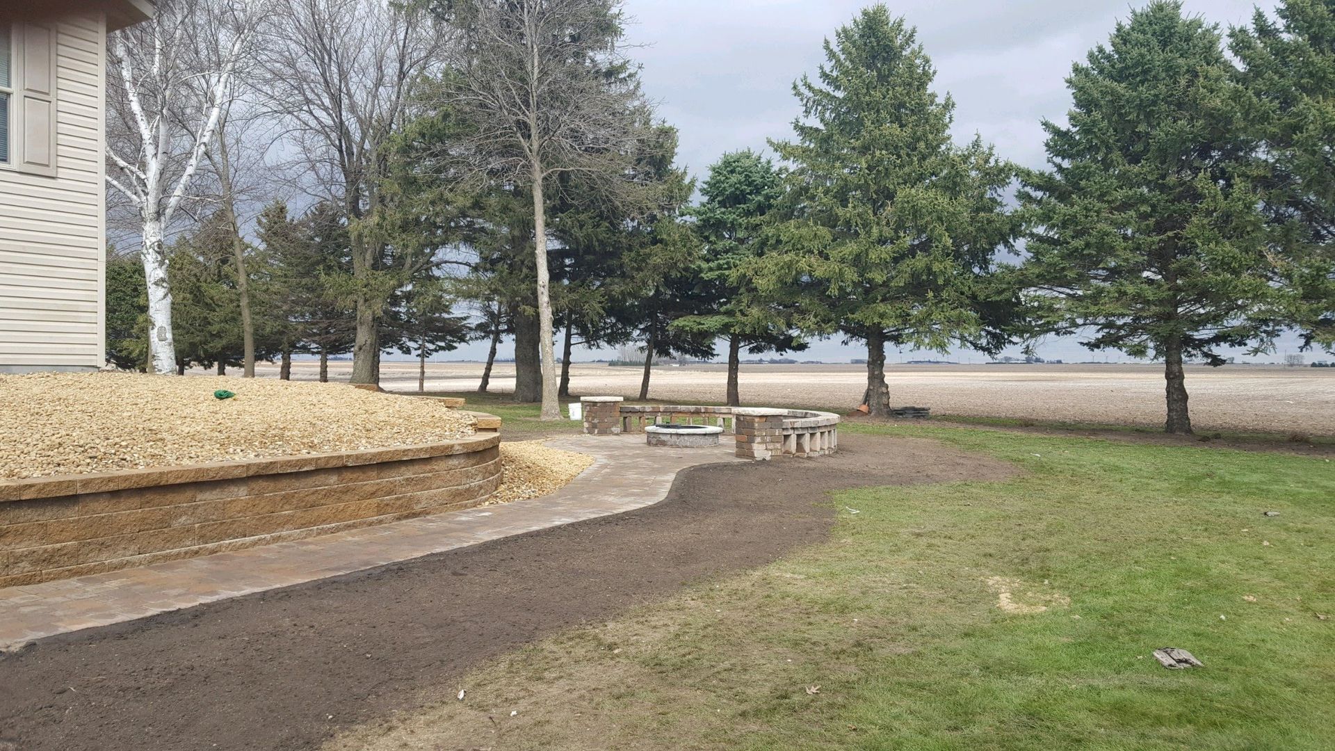 A brown stone retaining wall borders a dark, paved path leading to a picnic table in a yard with trees and a sandy shore.
