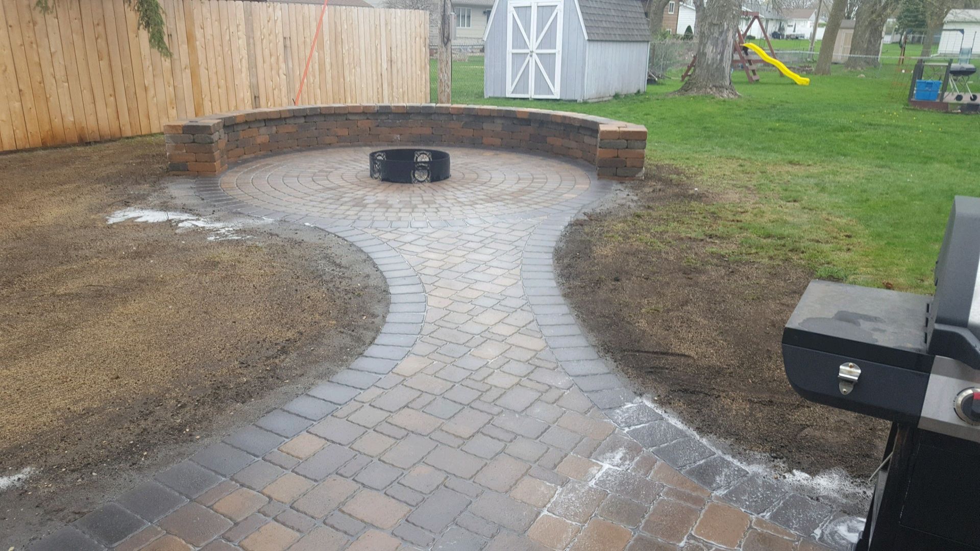 A paver patio with a curved stone seating wall and central fire pit, viewed from a grassy backyard.