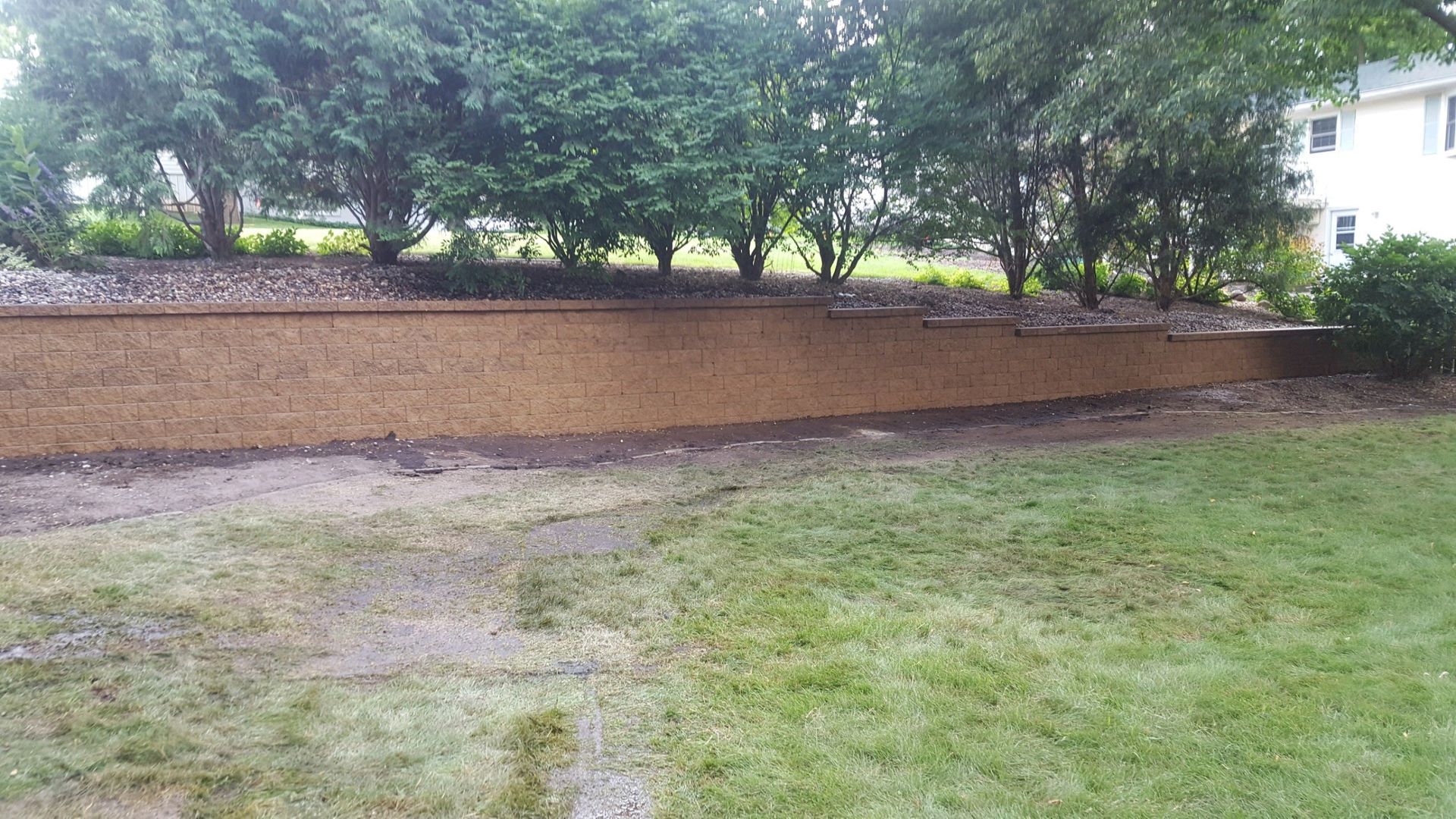 A tan, tiered retaining wall runs across a backyard, backed by a row of mature green trees and a white house.