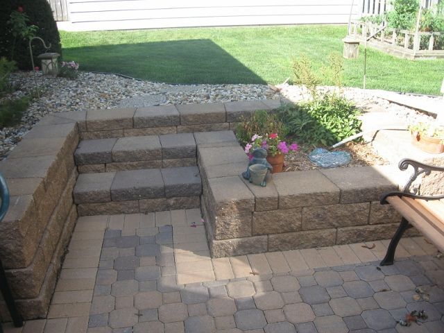 A stone patio with a built-in block wall and steps leading to a grass lawn, featuring a small garden area and a bench.