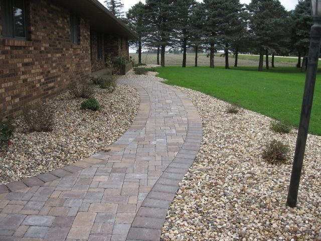 A winding paved walkway made of patterned bricks with a stone border, surrounded by gravel landscaping and a green lawn.