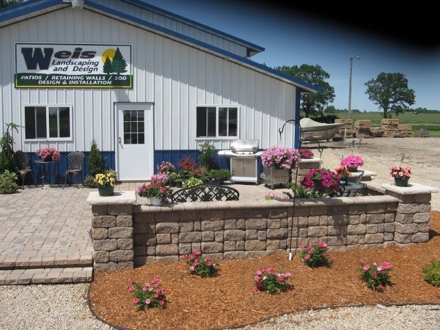 A landscape design showroom features a paver patio with a stone retaining wall, colorful potted flowers, and mulch beds.