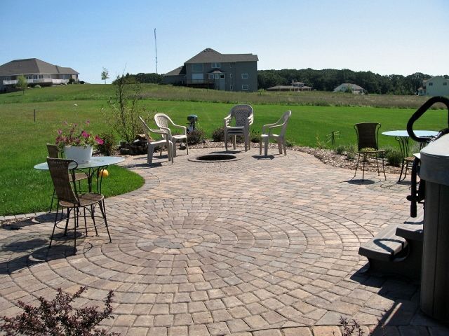 A circular stone patio with outdoor chairs and tables, featuring a fire pit, overlooking a large green grassy yard.