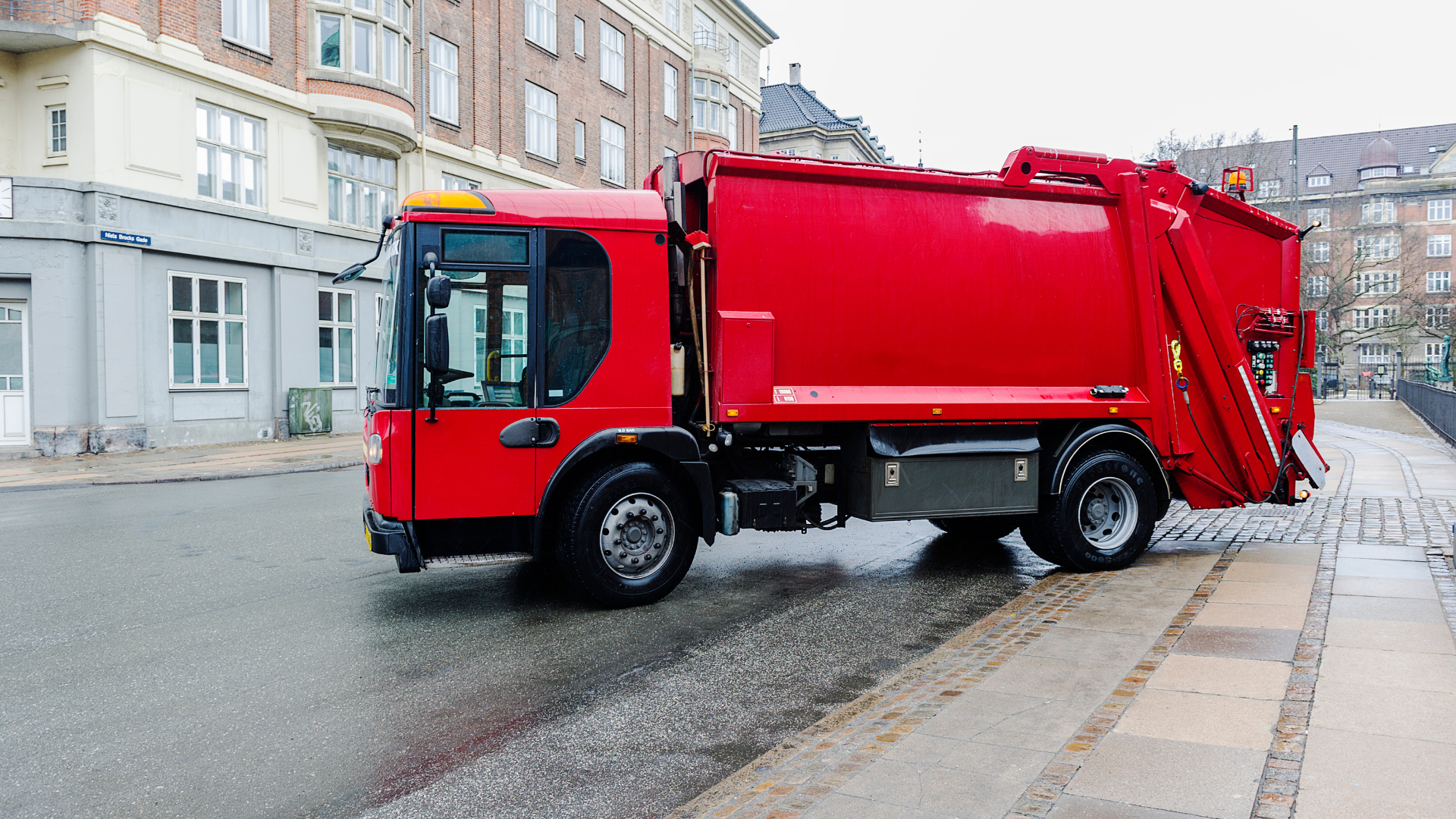 Red garbage truck on a wet street in front of buildings.