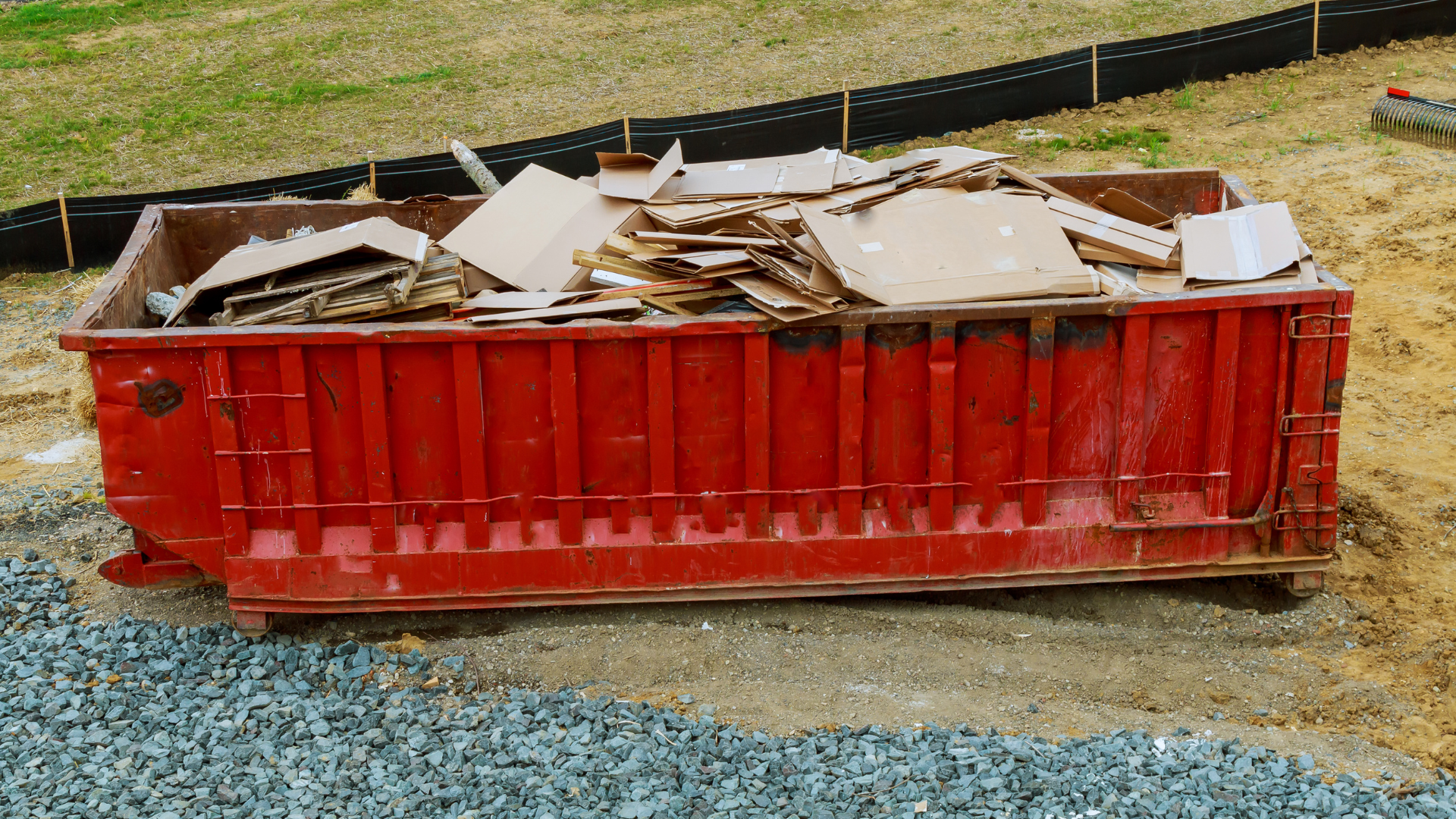 Red dumpster filled with construction debris on a gravel surface near a fence