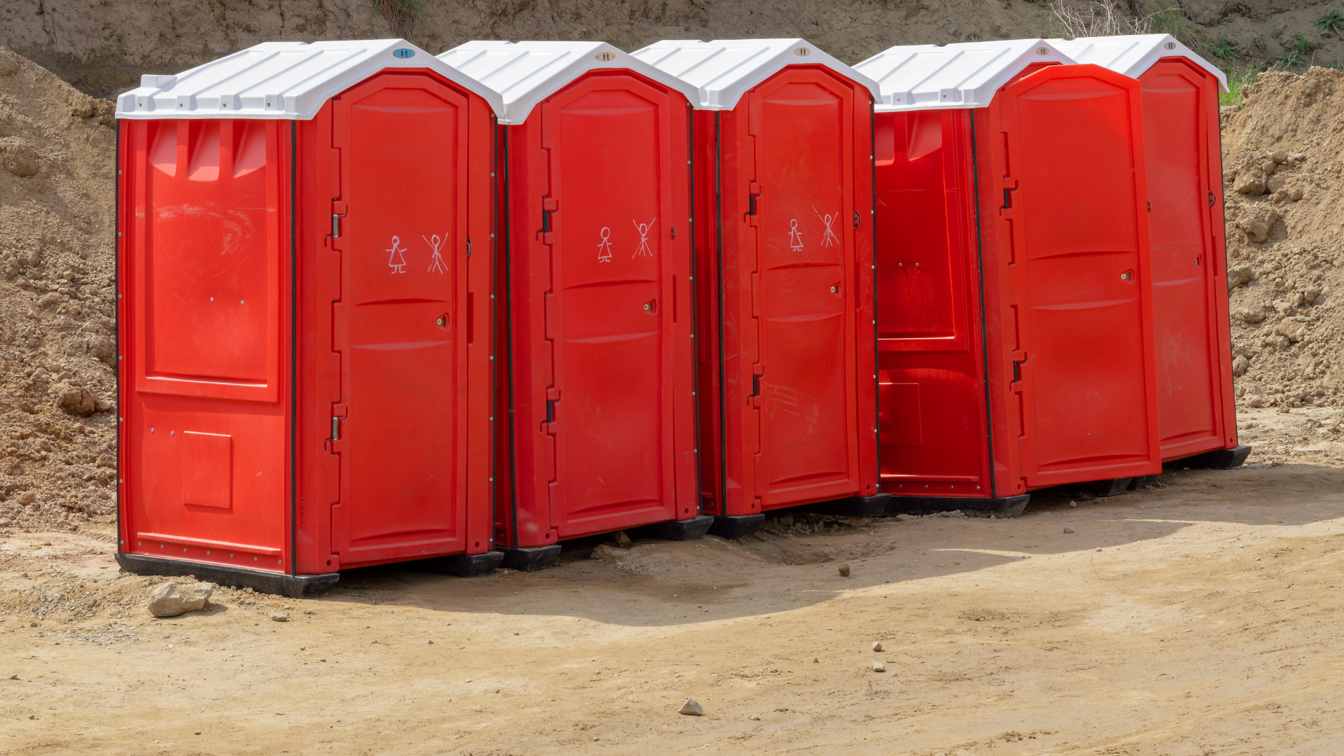 Red portable toilets in a row on dirt, with white roofs.