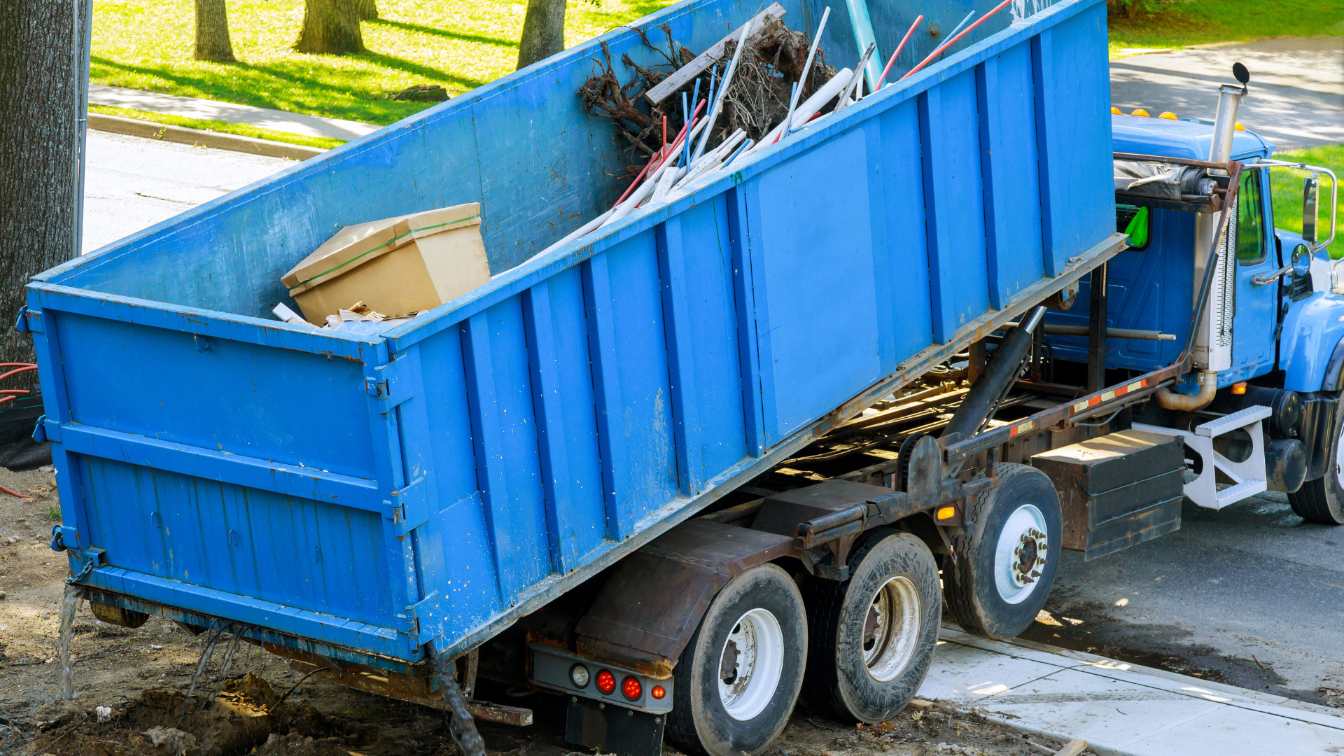 Blue dumpster being emptied by a truck, filled with debris, outdoors.