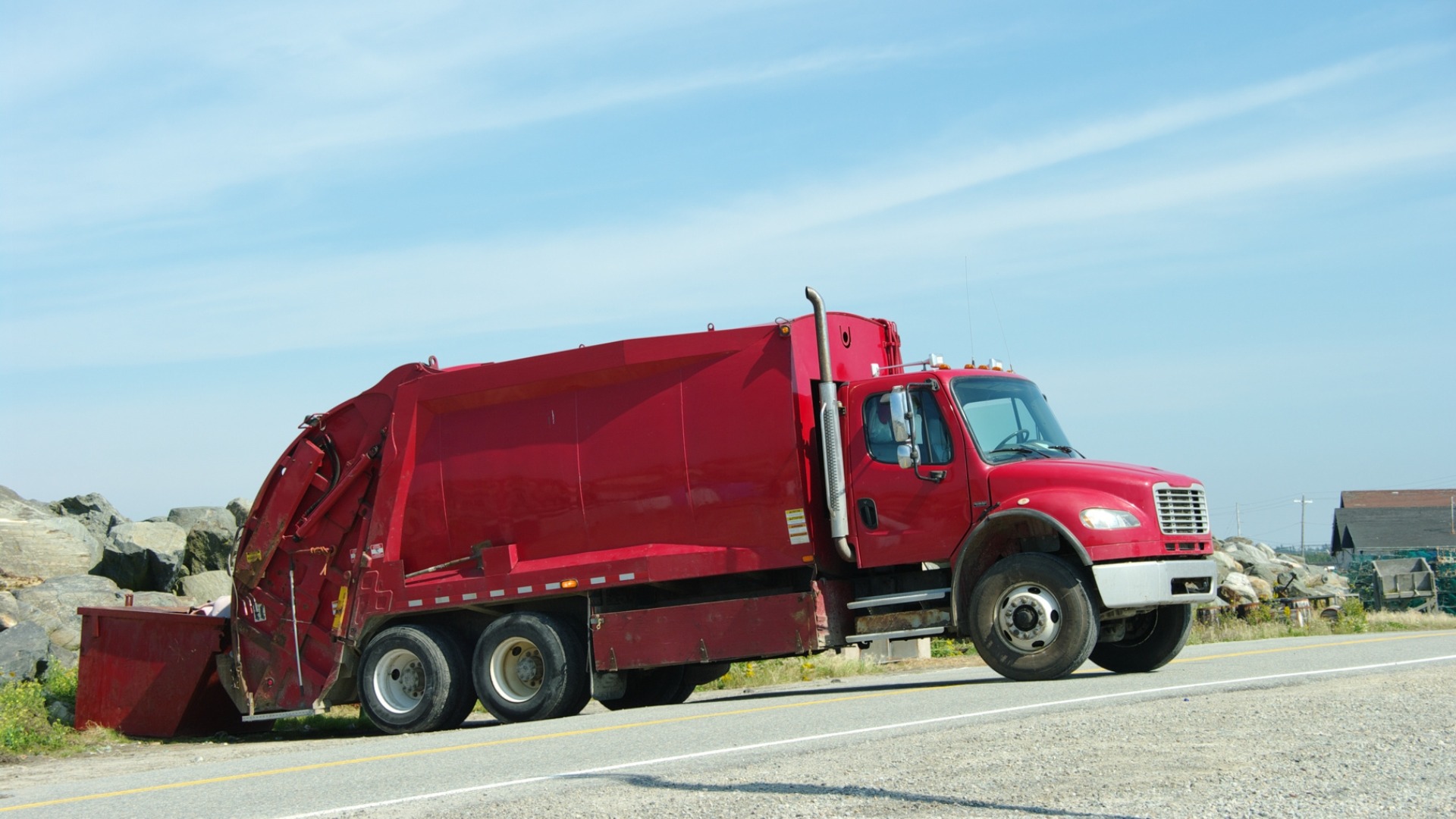 Red garbage truck on a road, driving towards the right, under a blue sky.