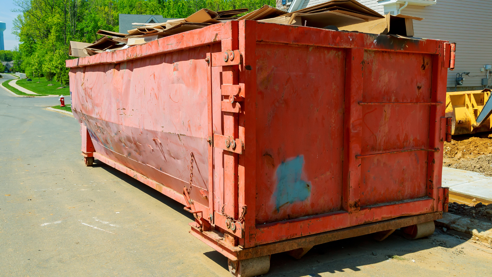 Red dumpster filled with debris, parked on a street near a construction site.