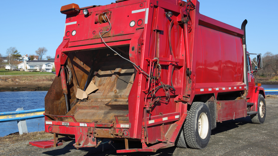 Red garbage truck, rear view, open bin, waterfront background.