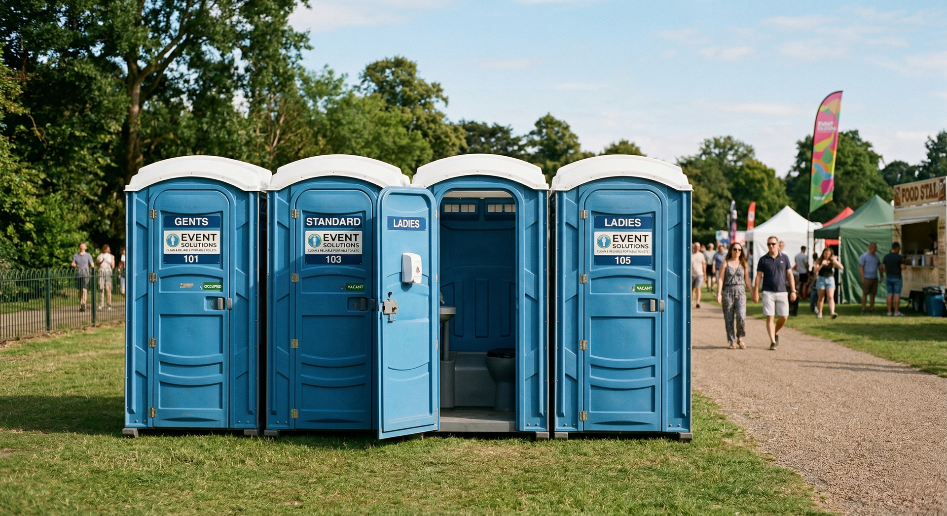 A row of four blue portable toilets stands on a grassy field at an outdoor event, with one door standing open.