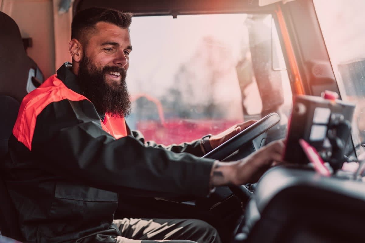 Truck driver with a beard smiling, wearing a safety vest, driving a truck.