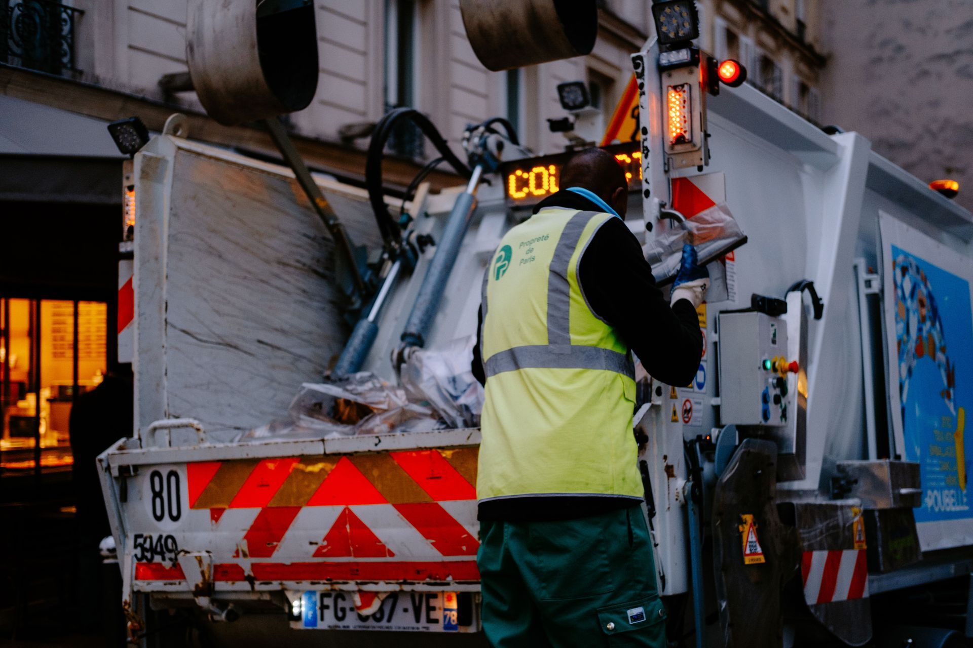 Sanitation worker in yellow vest operates a garbage truck at dusk, rear lift raised.