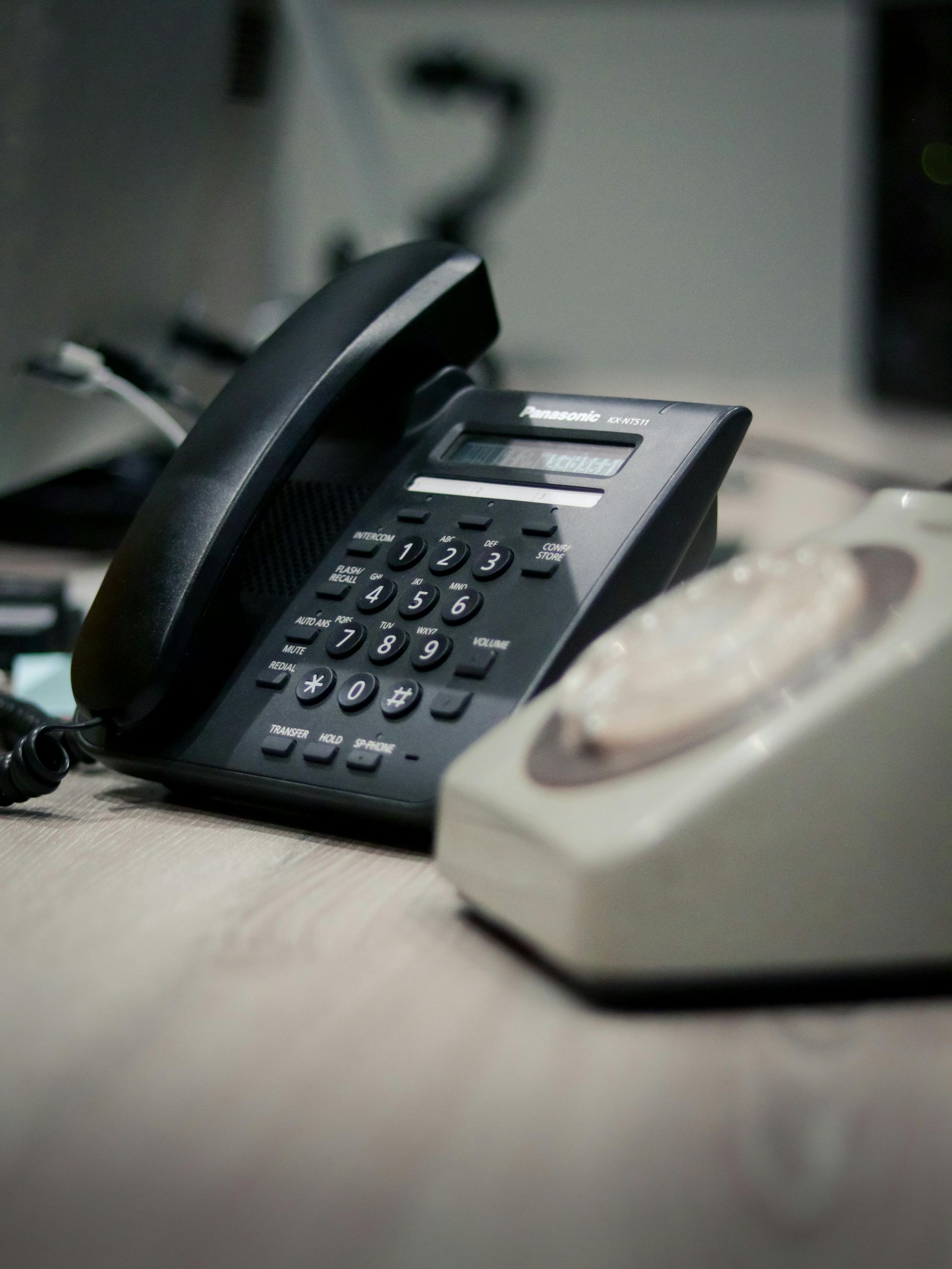 A modern black office telephone sits next to an older, beige rotary dial phone on a wooden desk.