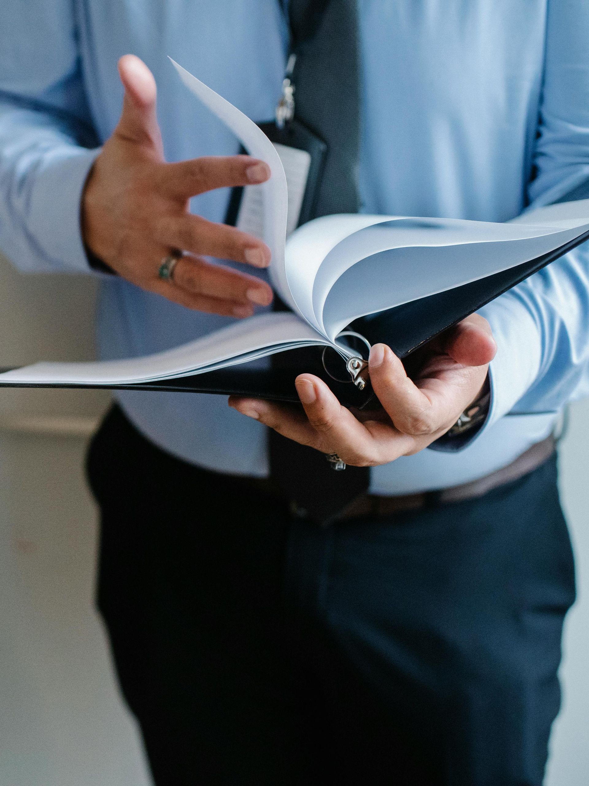 A person in a blue shirt and dark trousers holding and flipping through the pages of an open black binder.