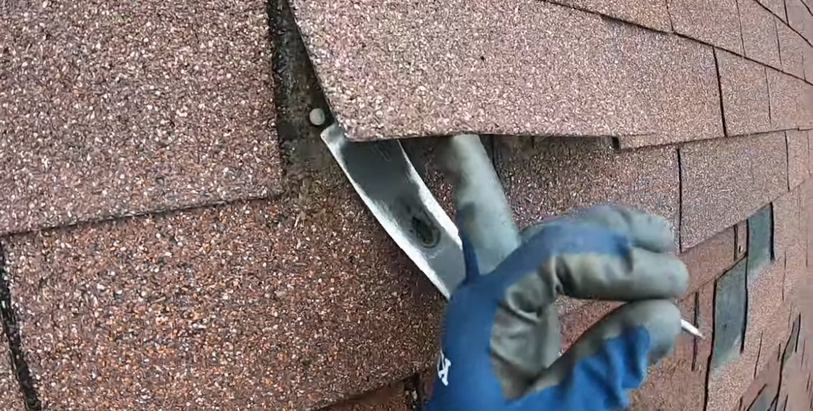 Gloved hand lifting a brown roofing shingle to show a metal flashing.