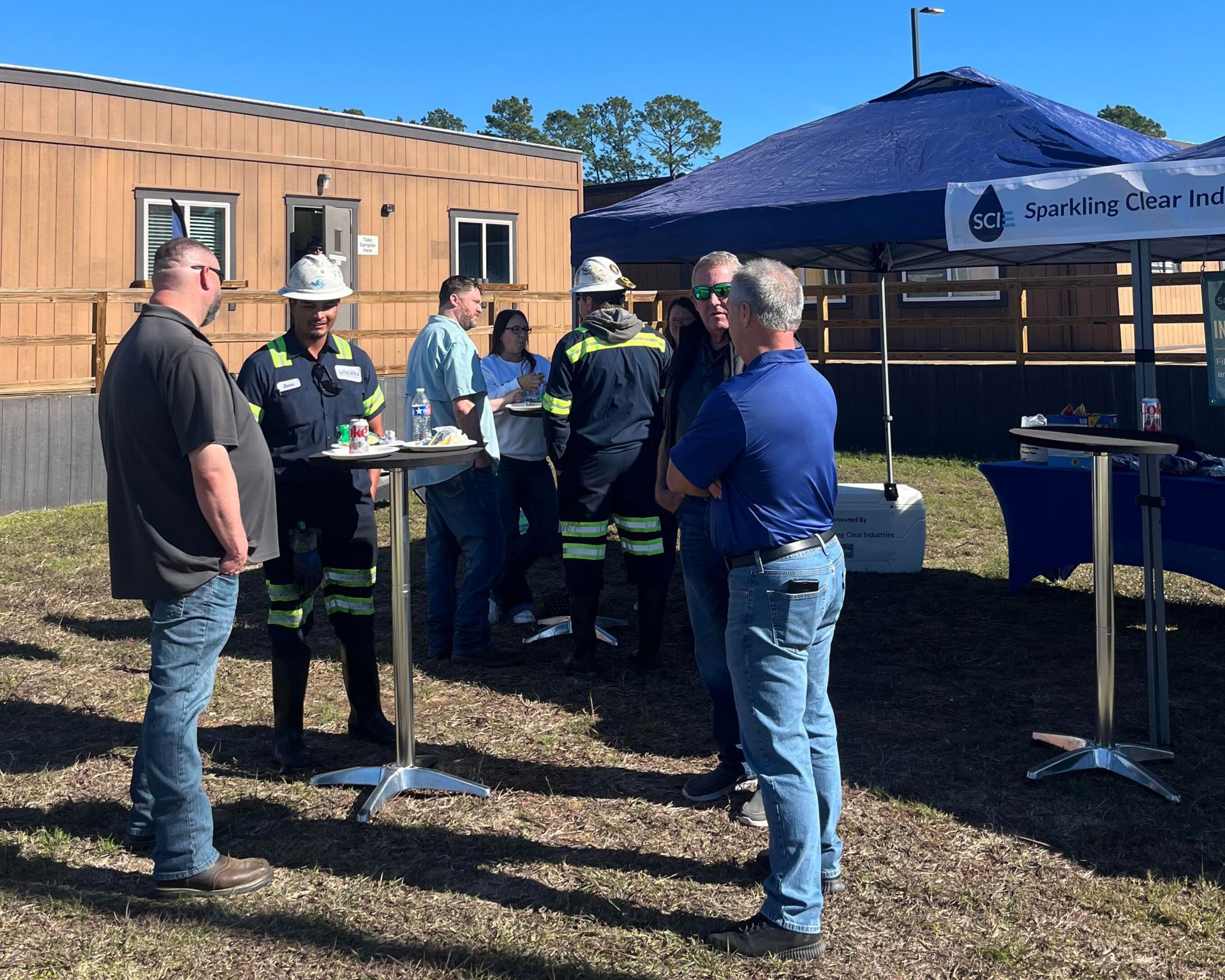 People in work attire and hard hats at an outdoor event near a tent and a building.
