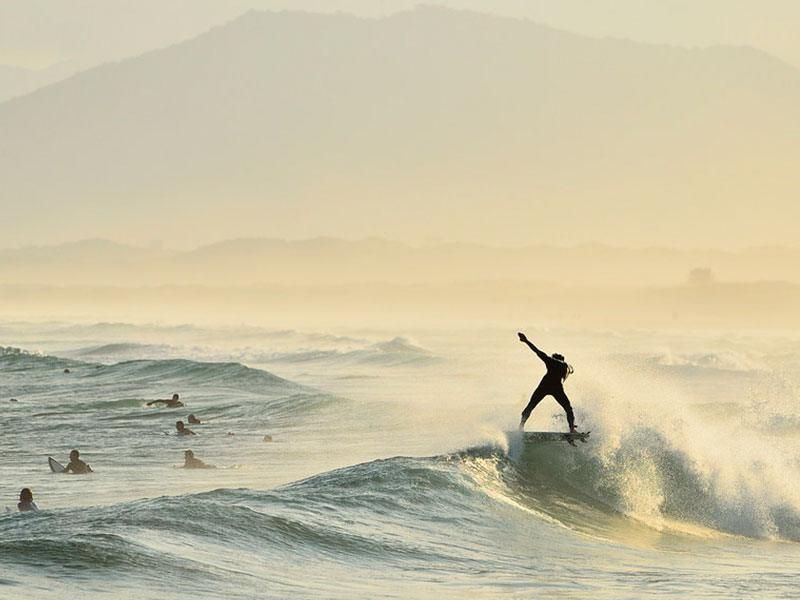 Surfista deslizando sobre uma onda, braços erguidos em sinal de triunfo. Cena de praia envolta em névoa, com outros surfistas ao fundo e uma montanha.