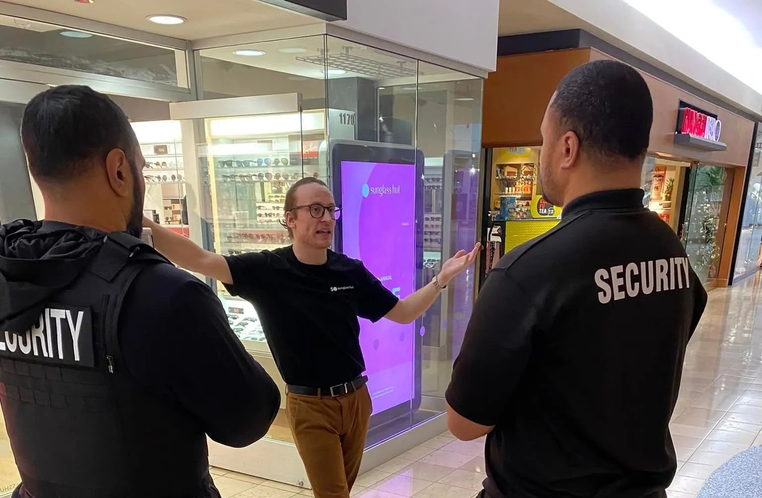 Three security guards speaking in a mall corridor, one gesturing toward a storefront.