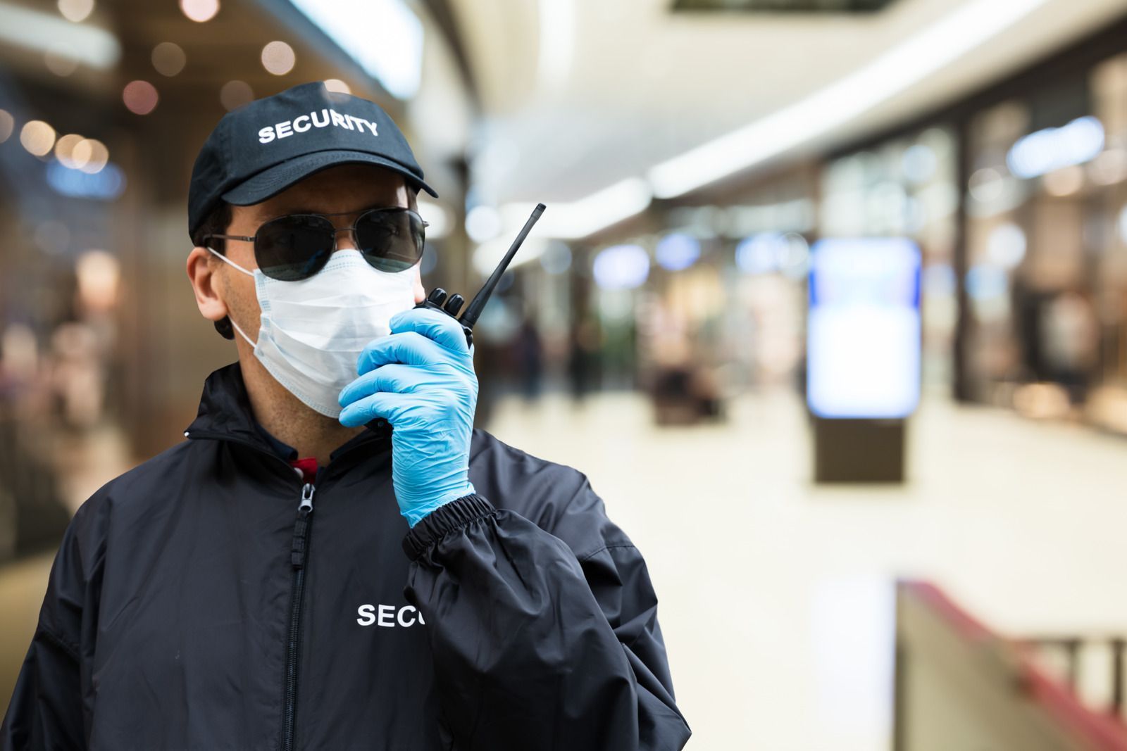 Security guard in black uniform and blue gloves wearing a mask in a blurred indoor hallway