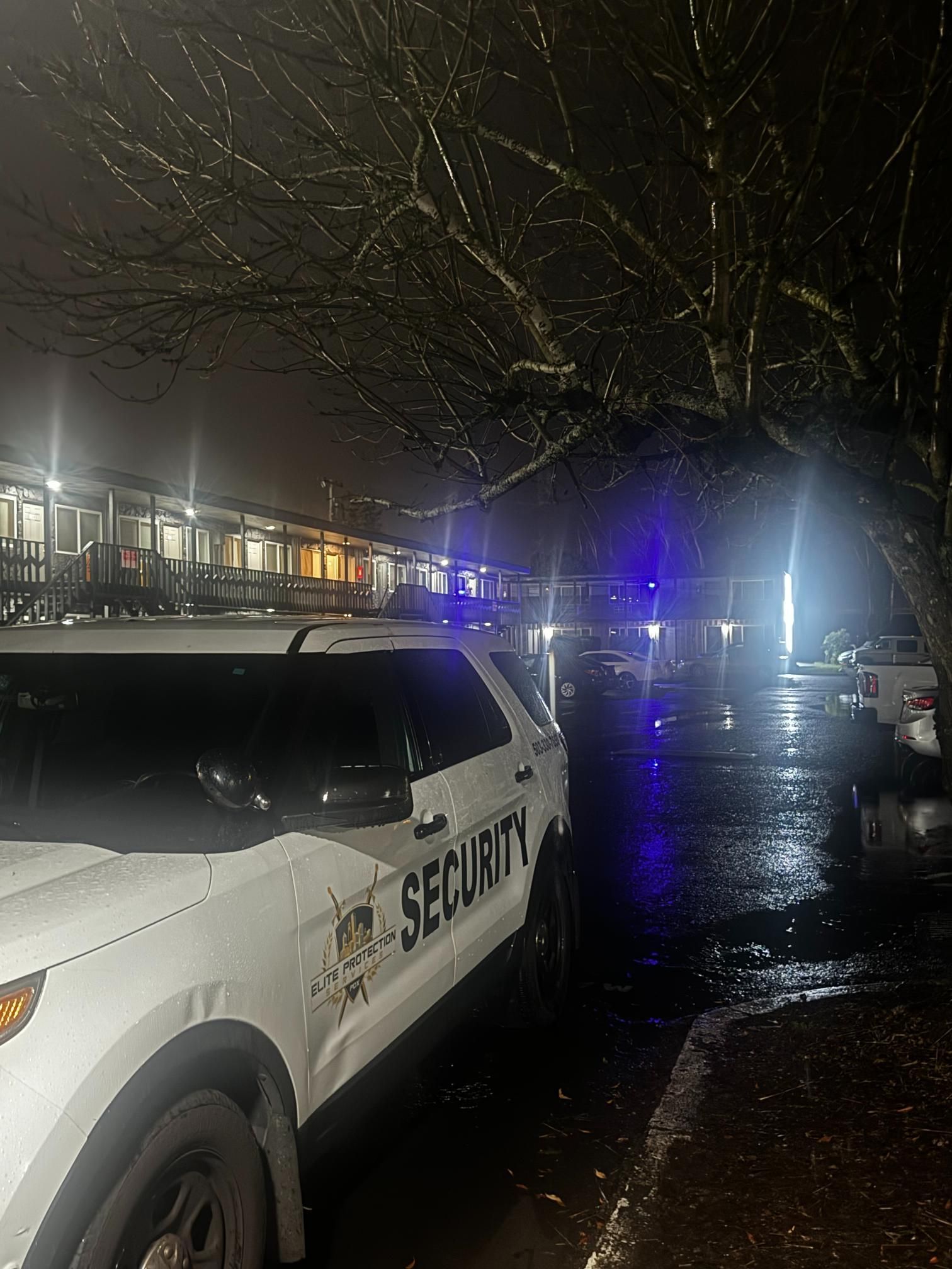 Security SUV parked at night near flashing blue lights outside a wet, tree-lined building
