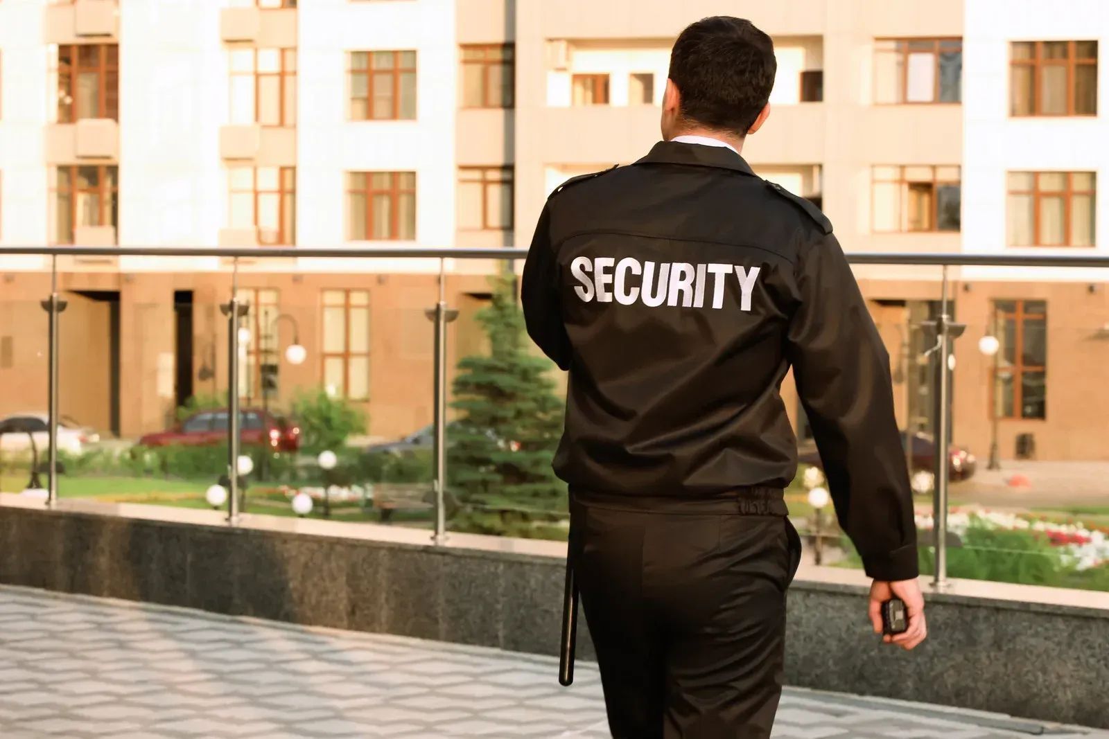 Security guard walking outside a building, seen from behind, wearing a black jacket with “SECURITY” on the back.