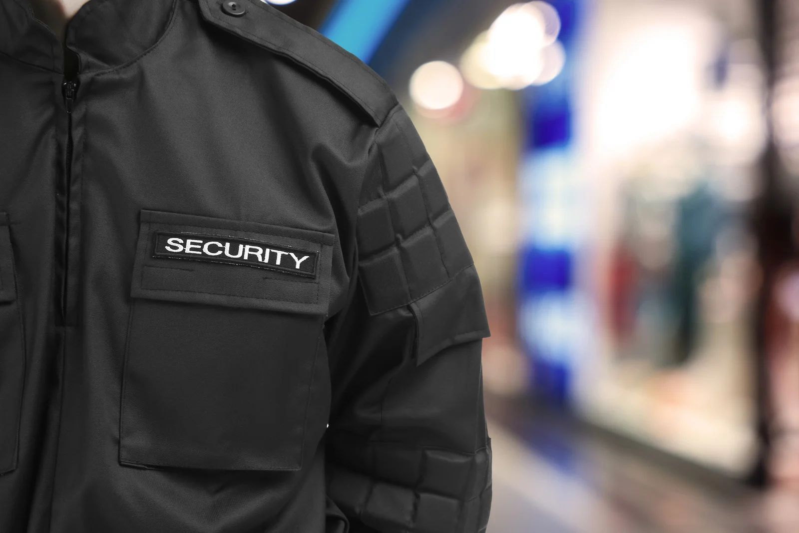 Security guard in black jacket with “SECURITY” patch in a blurred indoor hallway