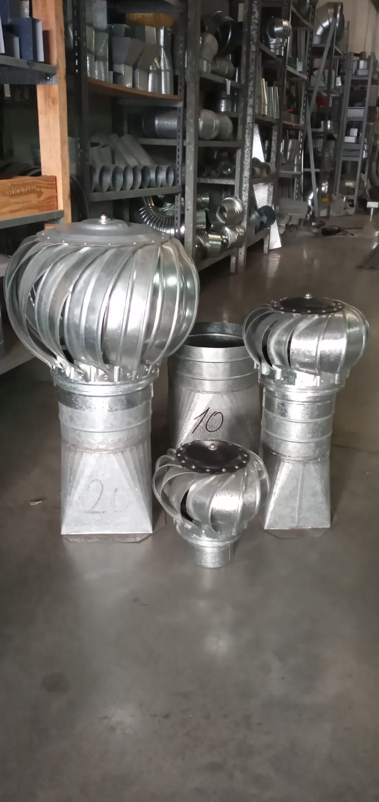 Tres ventiladores de techo de turbina de metal plateado expuestos en el suelo de un almacén frente a unas estanterías.
