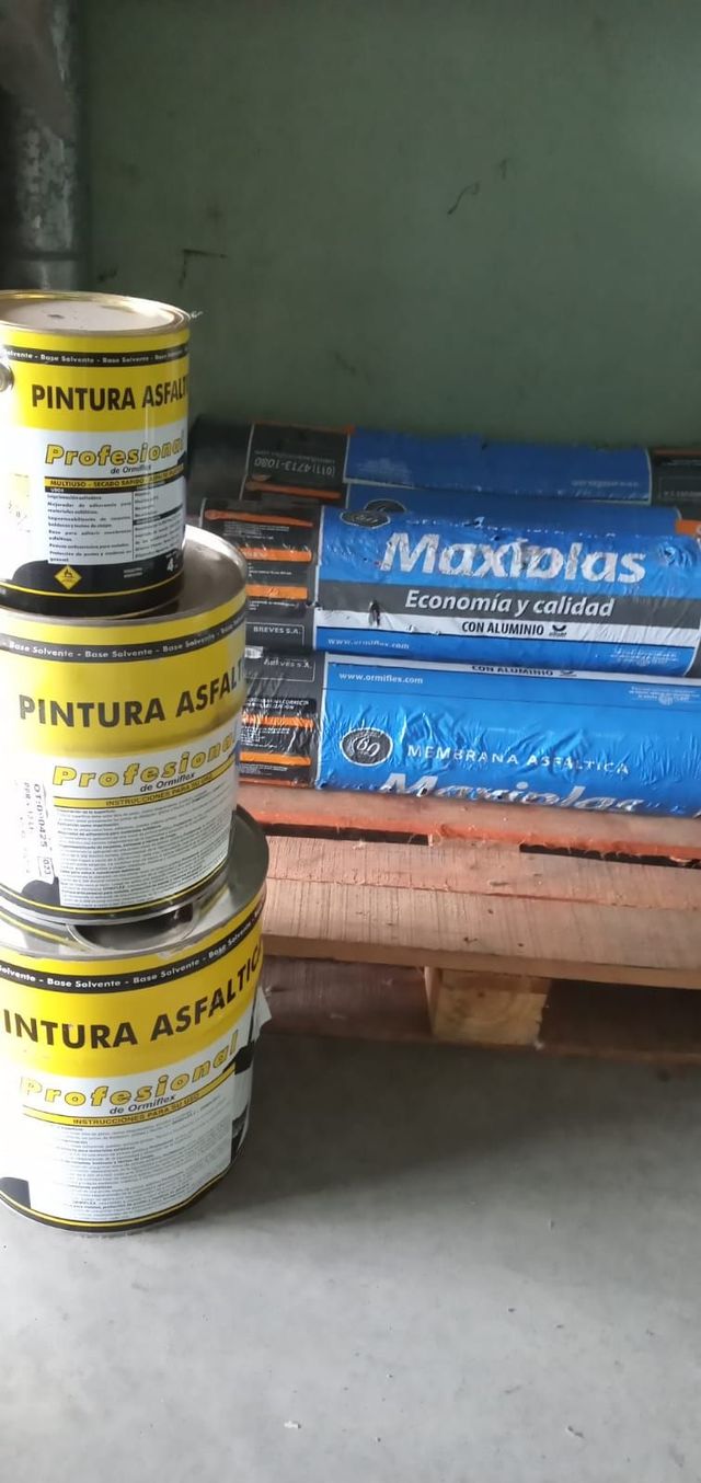 Pilas de latas de pintura amarillas y rollos de material azul sobre palés de madera en un almacén.