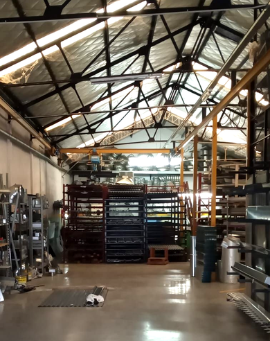 Interior de un almacén con estanterías metálicas, vigas altas y equipos dispersos en el suelo.