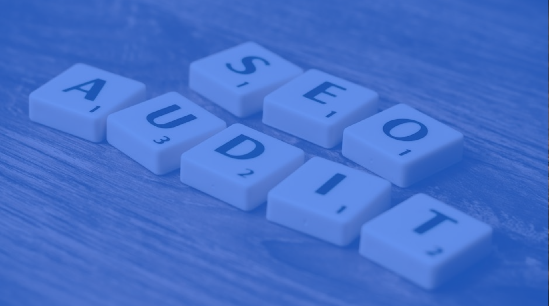 Blue-tinted Scrabble tiles spelling “AUDIT” on a wooden surface.