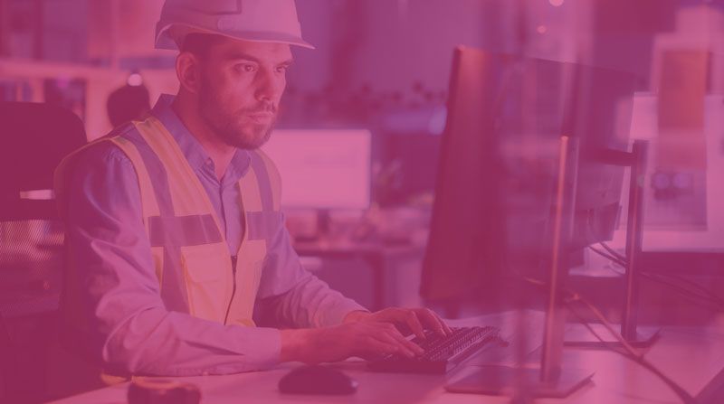 Worker in hard hat and safety vest typing at a computer in a pink-lit office.