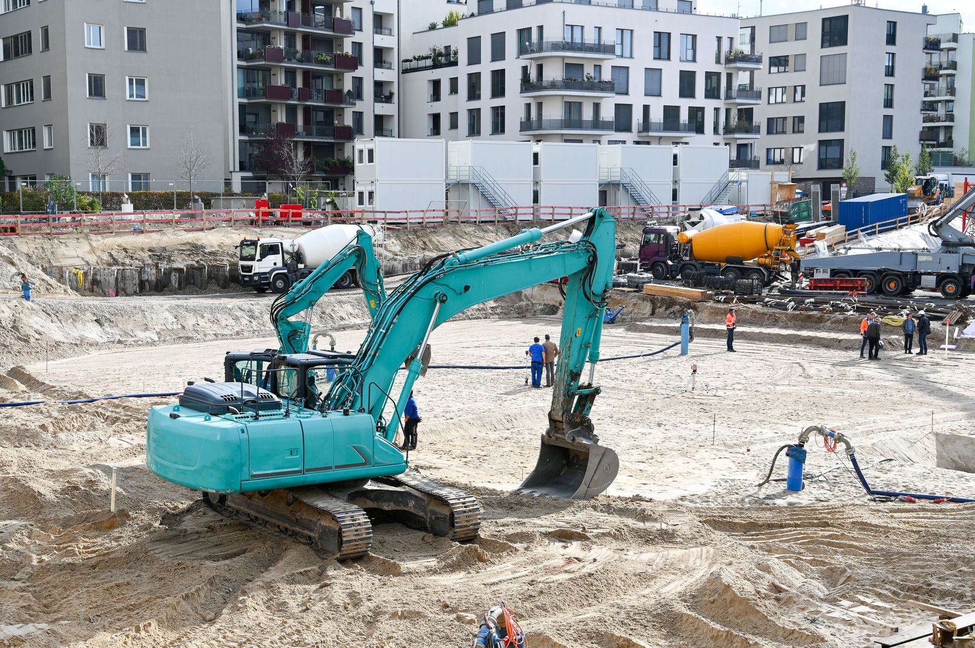 Turquoise excavator digging at a construction site with workers and apartment buildings in the background