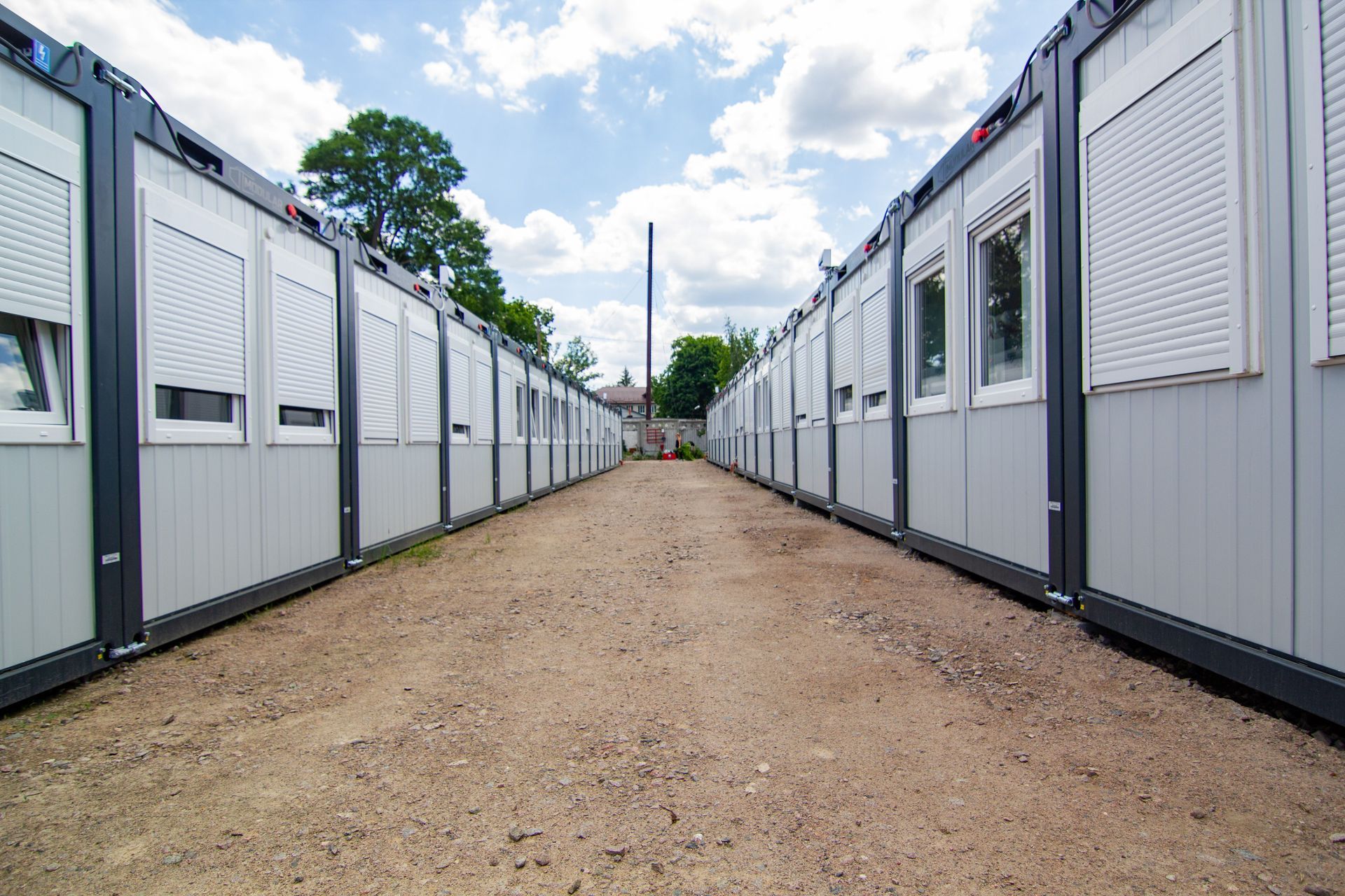 Row of gray portable buildings lining a gravel path under a partly cloudy sky