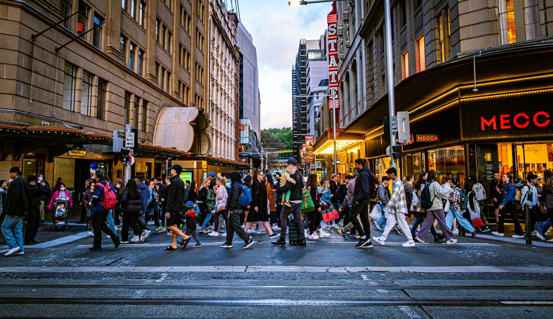 Crowded city crosswalk in Sydney at dusk with pedestrians, tall buildings, and bright storefront lights.