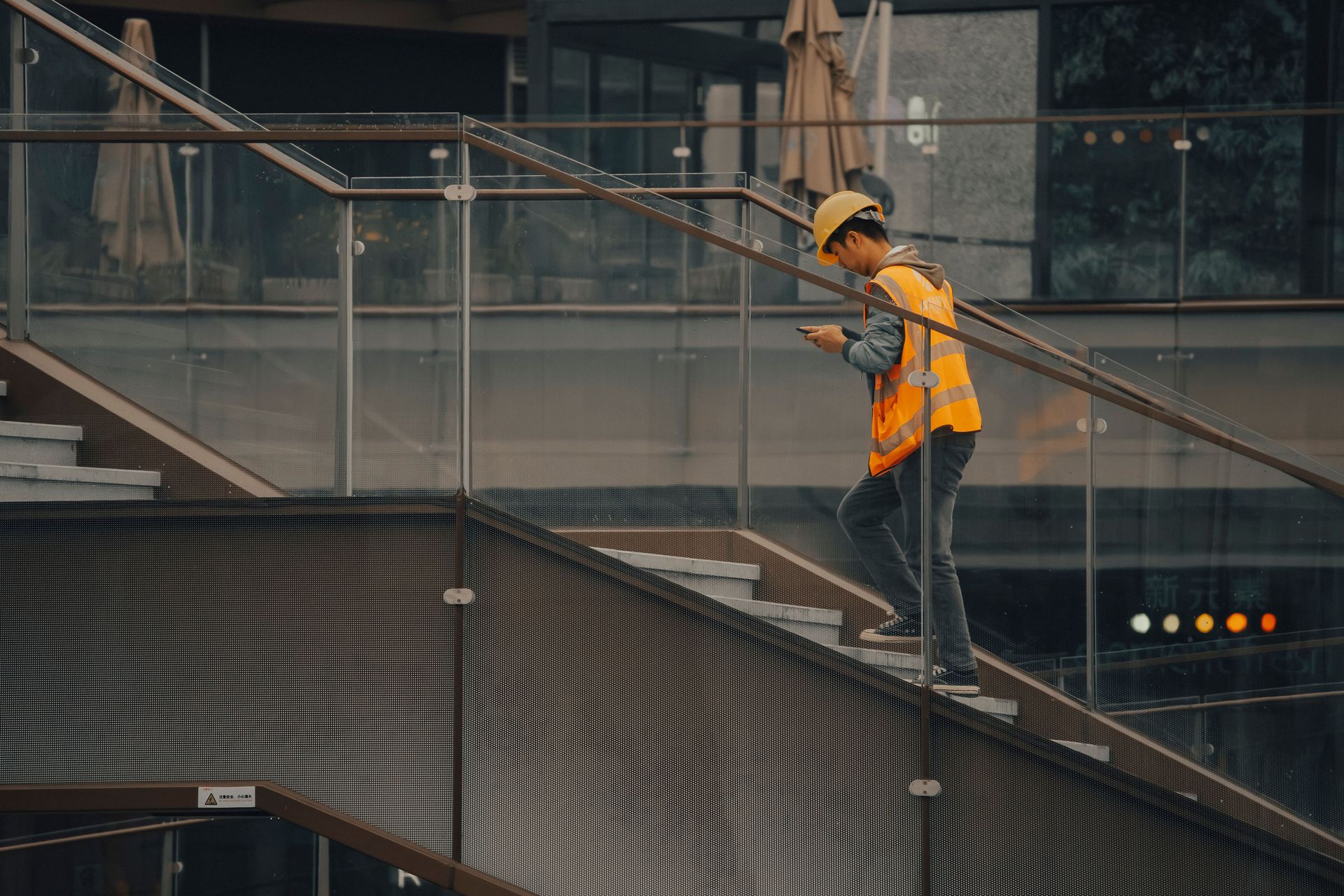 Construction worker in an orange safety vest and hard hat on a metal staircase beside a building.