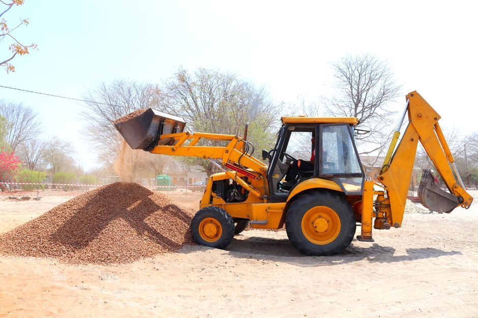 Yellow backhoe loader dumping dirt beside a pile on a dusty roadside construction site