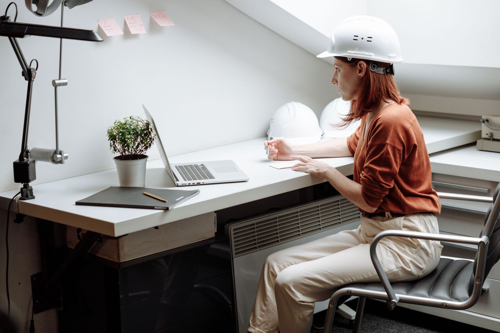 Person in a white hard hat working at a desk with a laptop in a bright attic office.