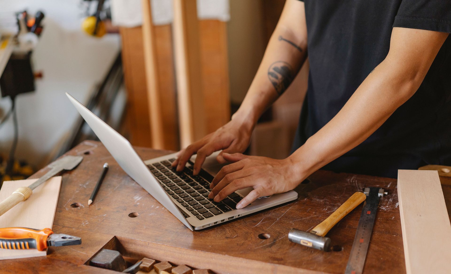 Person typing on a laptop at a wooden workbench with tools nearby