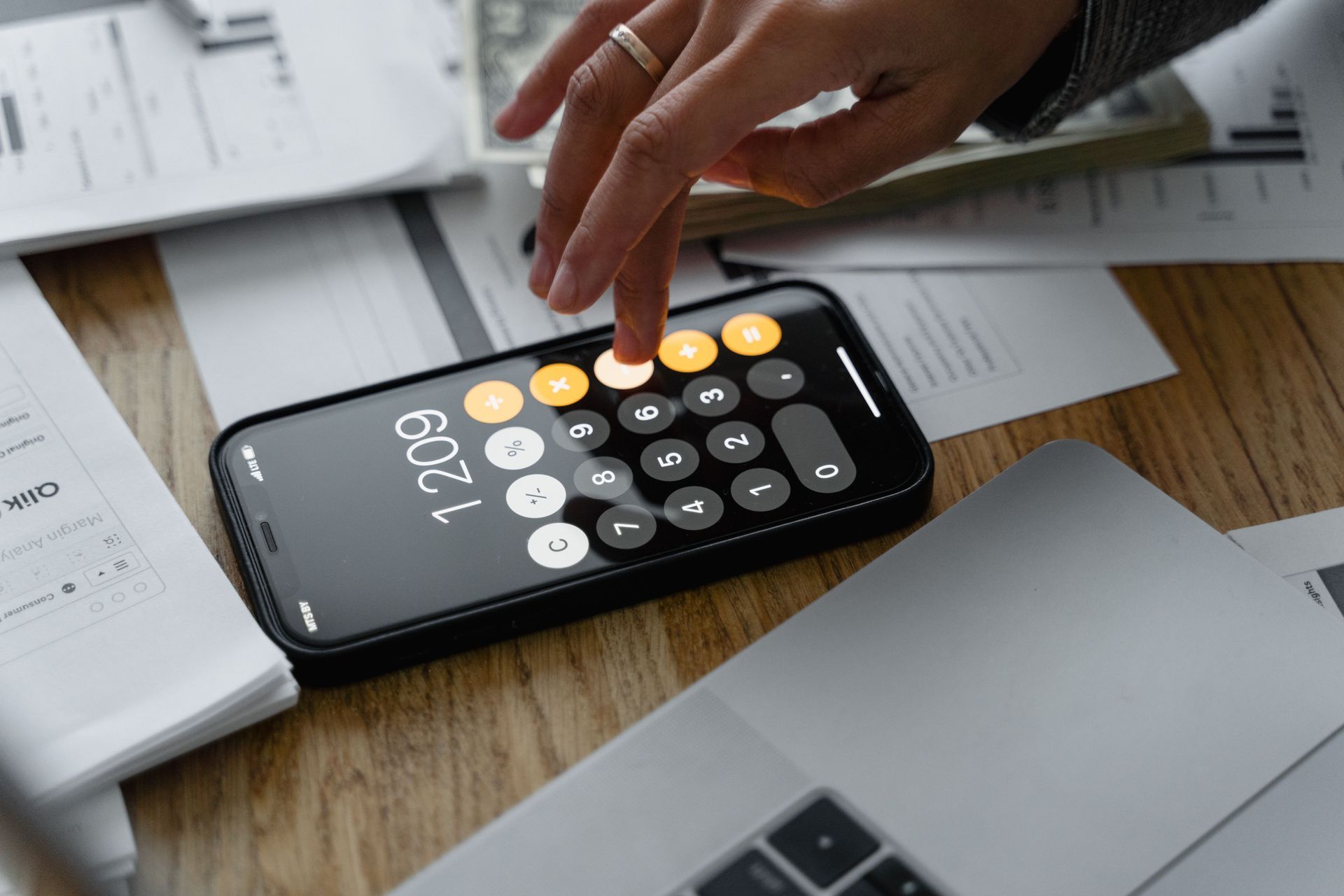 Hand tapping a calculator phone on a desk beside papers and a laptop
